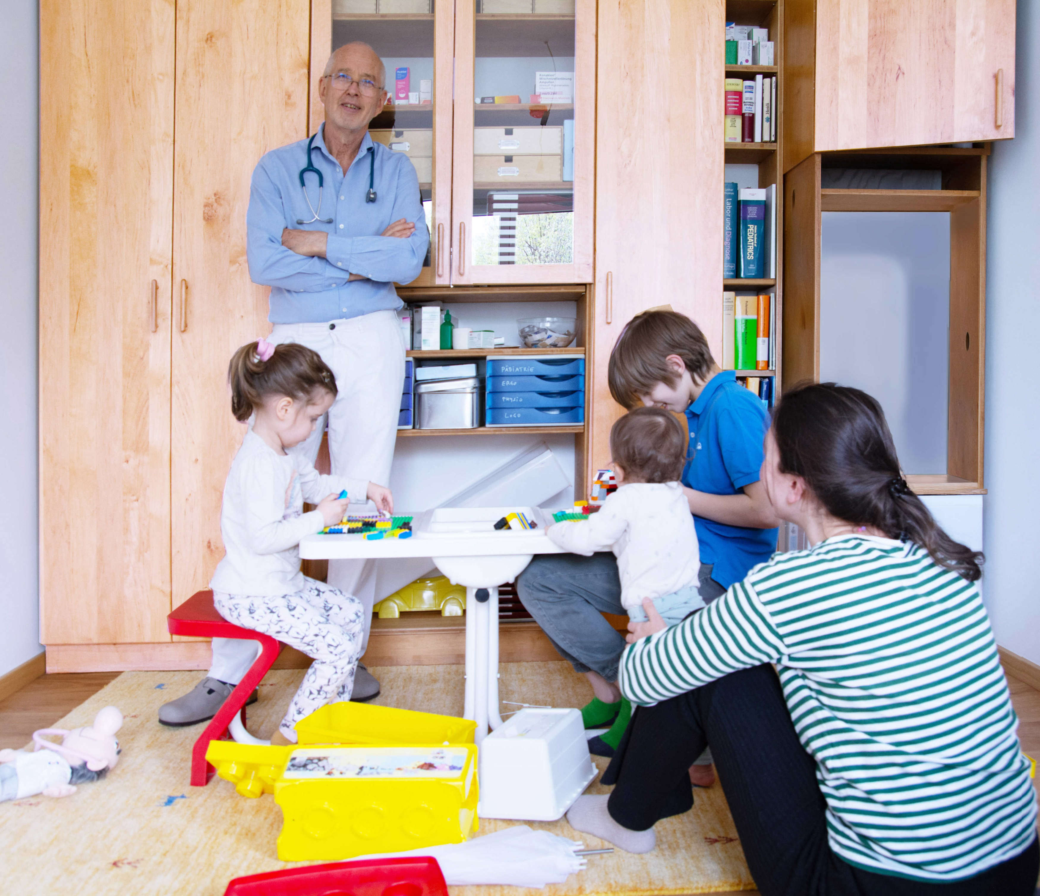 In der Ordination. Kinder spielen an einem Tisch. Mutter sitzt davor. Kinderarzt Dr. Pelzl steht daneben.